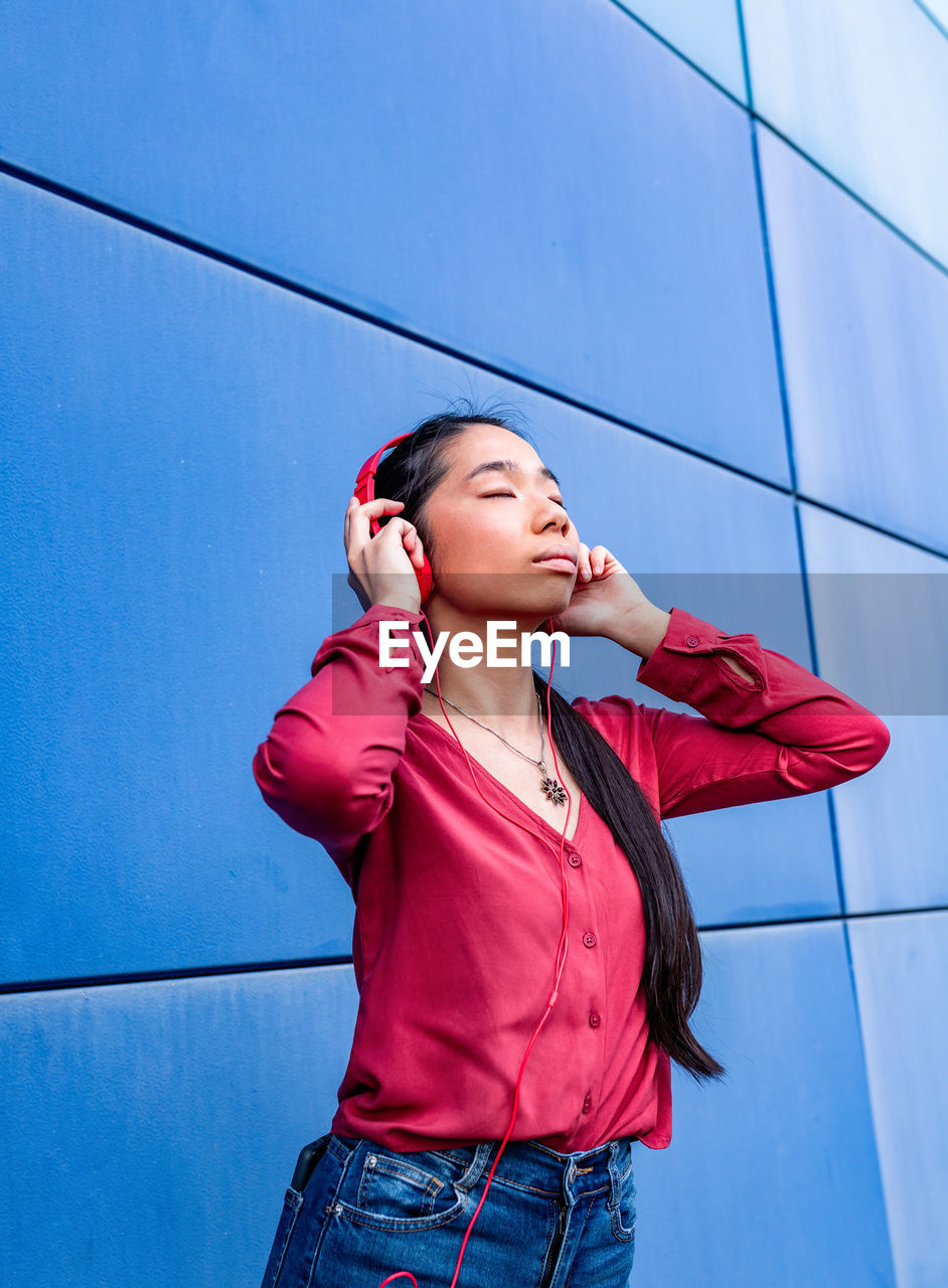 Ethnic female with closed eyes listening to music in headphones standing against blue wall
