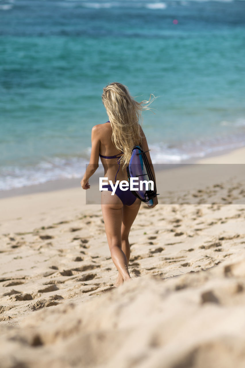 Rear view of woman in bikini walking with surfboard at beach
