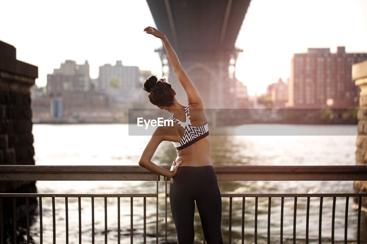 Rear view of sporty woman stretching by railing under williamsburg bridge