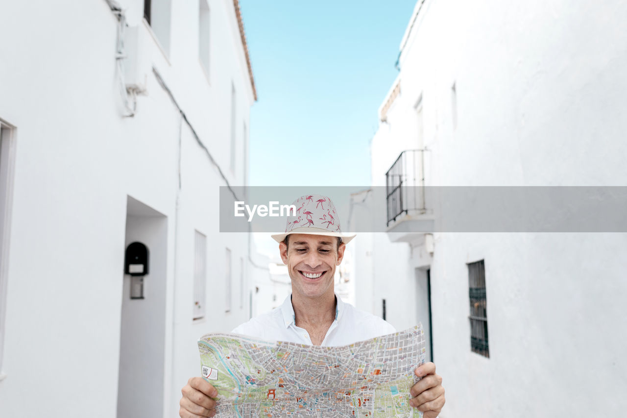 Content male tourist in ornamental hat standing with map between aged houses in white village in rhodes