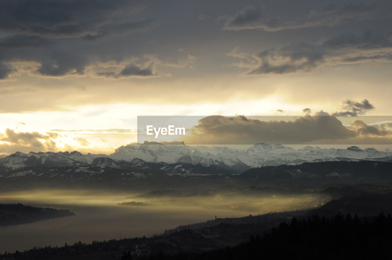 Scenic view of lake zurich by snowcapped mountains against cloudy sky during sunset