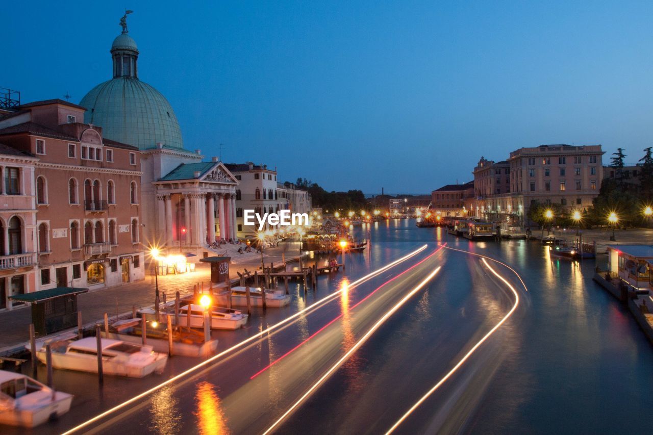 ILLUMINATED STREET AMIDST BUILDINGS AGAINST SKY AT DUSK