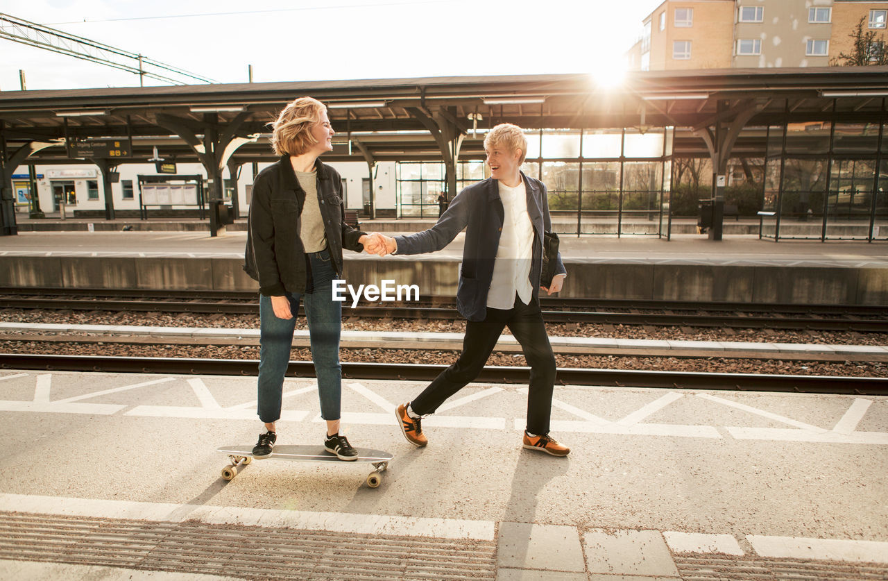 Cheerful young man pulling teenage girl on skateboard at railroad station platform