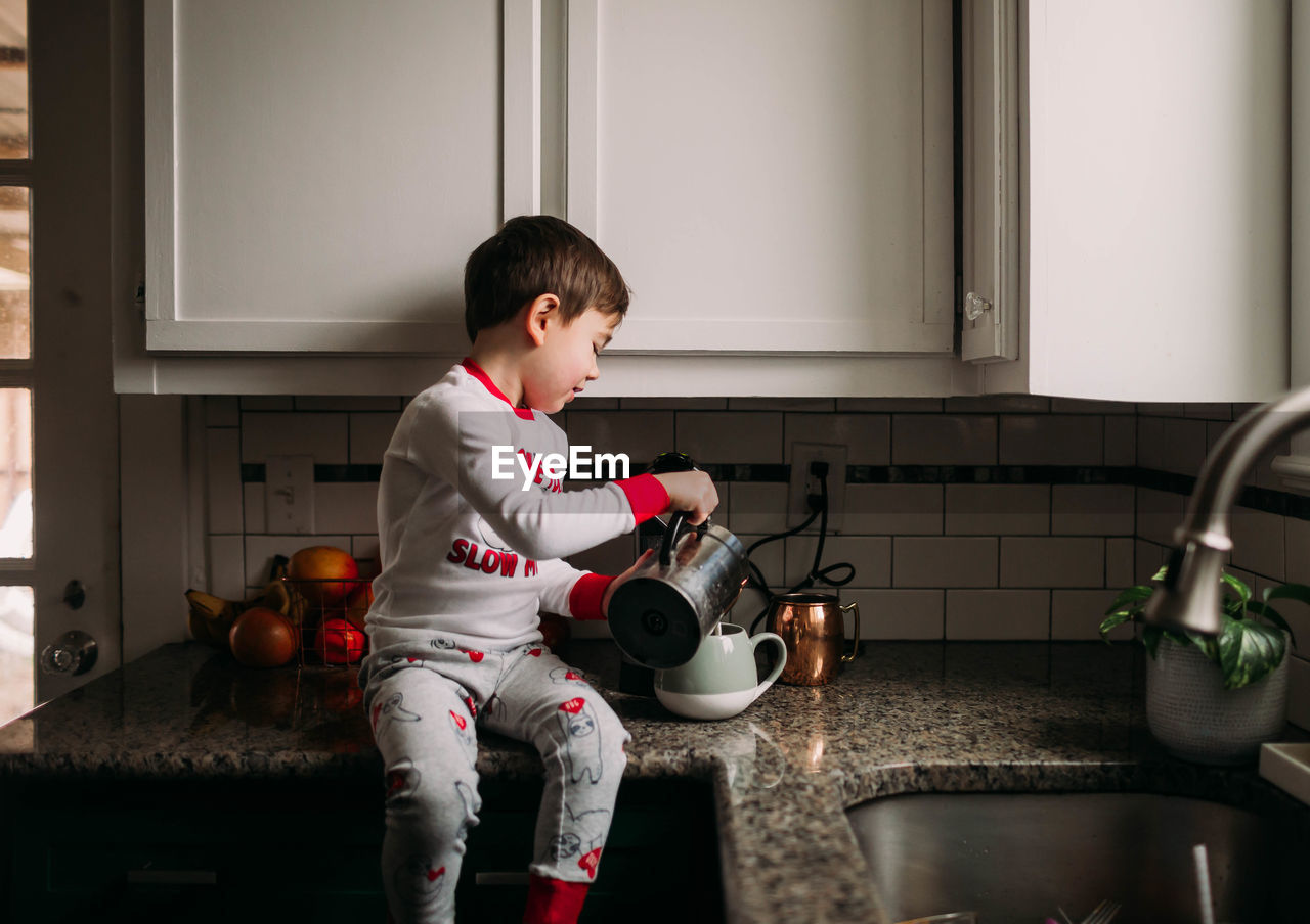 Young boy sitting on kitchen counter pouring milk into coffee