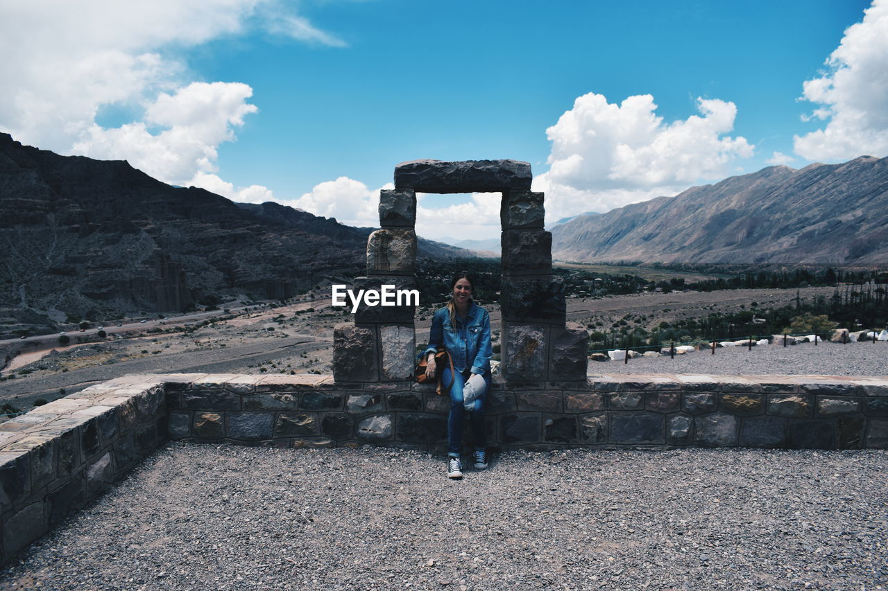 Portrait of woman sitting on retaining wall by mountains against sky