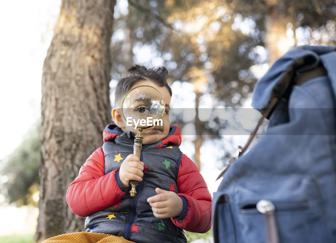 Boy in padded jacket looking through magnifying glass while sitting in park