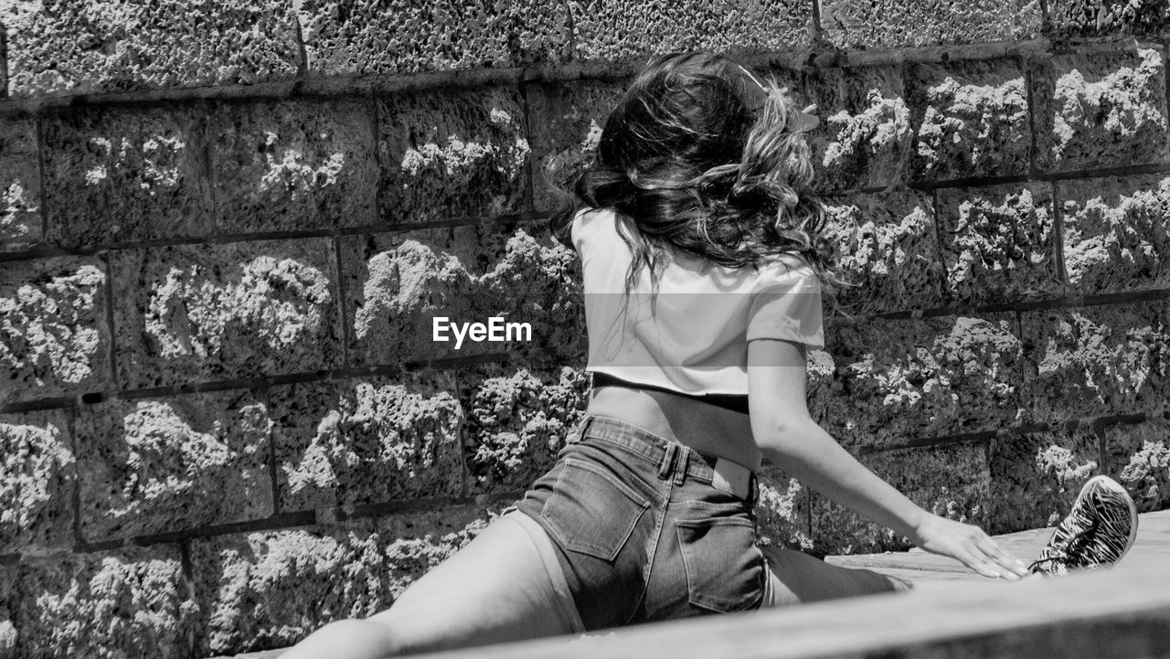 REAR VIEW OF WOMAN SITTING ON RETAINING WALL AGAINST BUILDING