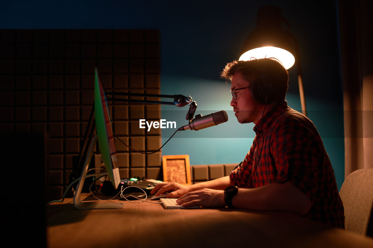 Side view of focused young male in checkered shirt and eyeglasses using computer and speaking in mic while recording podcast in dark studio