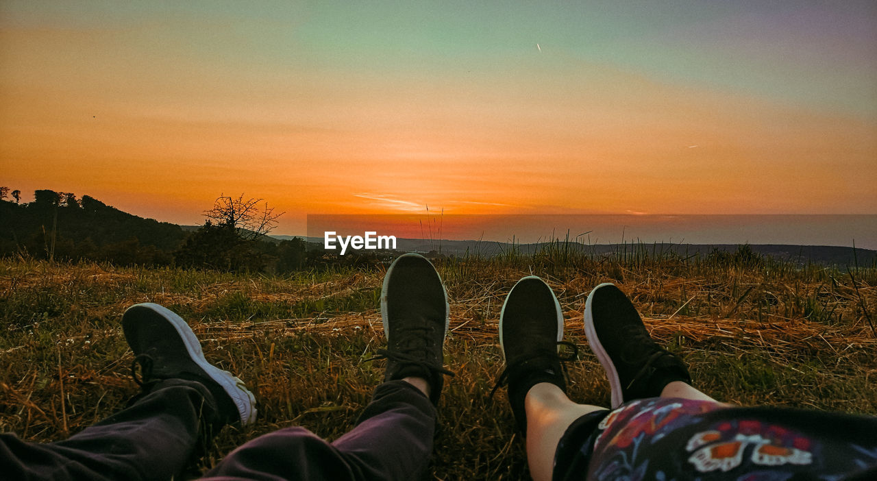 LOW SECTION OF MEN RELAXING ON LAND AGAINST SKY DURING SUNSET
