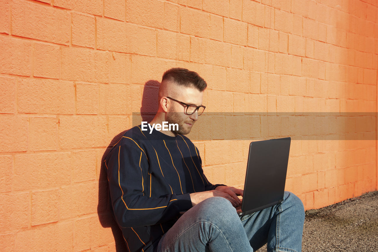 Man using laptop while sitting against brick wall