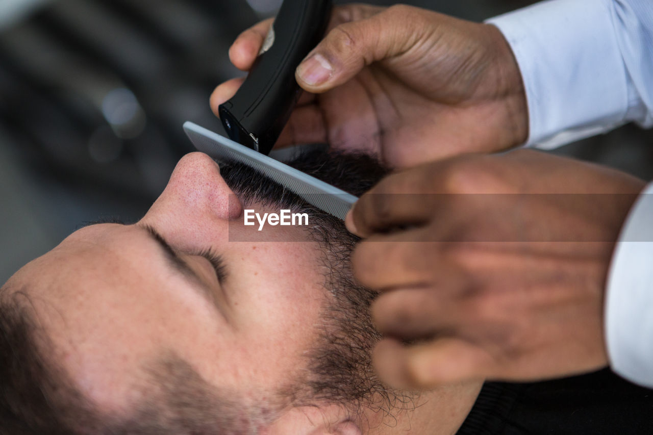 Crop anonymous ethnic male beauty master cutting mustache of bearded client using trimmer and comb in barbershop