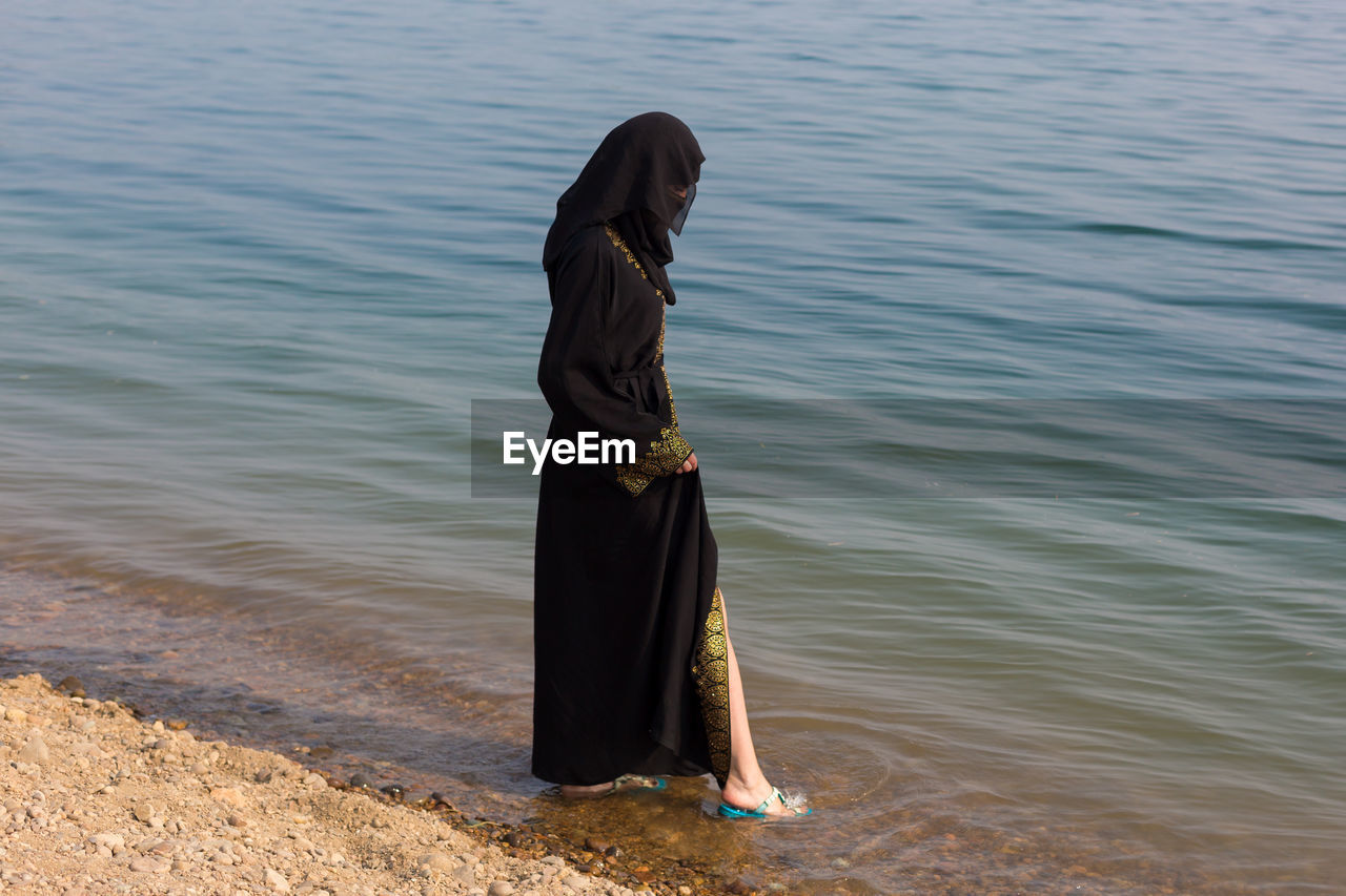 A muslim woman in national clothes wets her feet in sea.