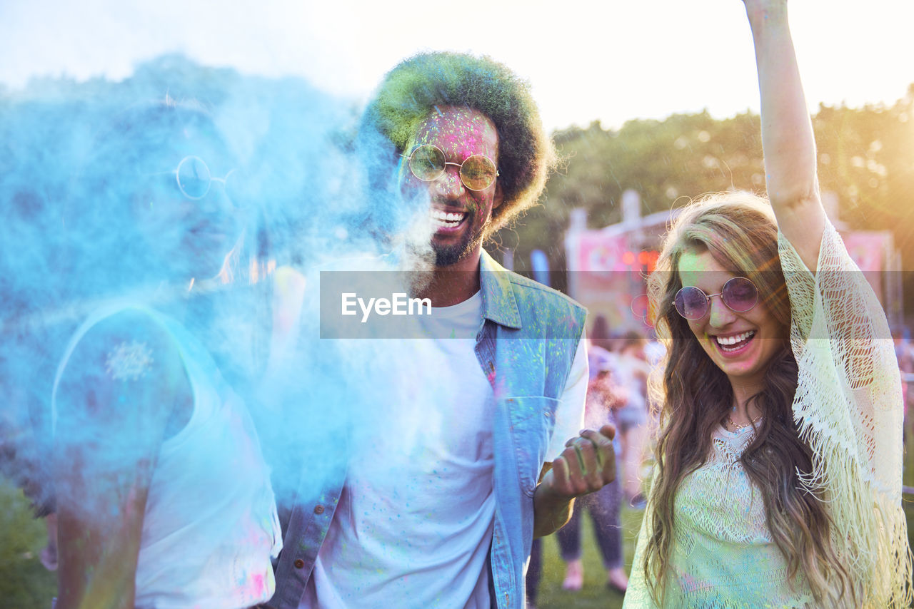 Portrait of smiling friends dancing while playing with powder paint against sky