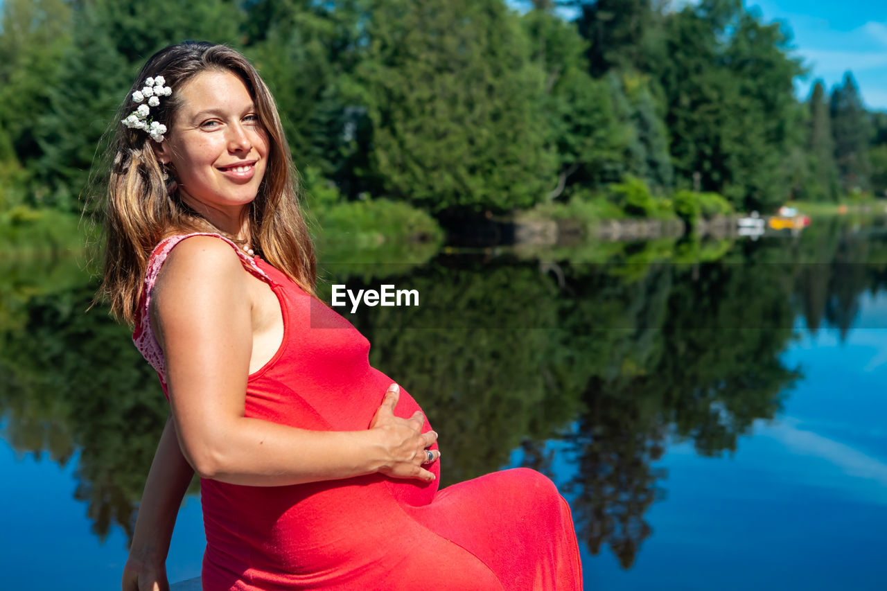 SMILING YOUNG WOMAN STANDING AGAINST LAKE