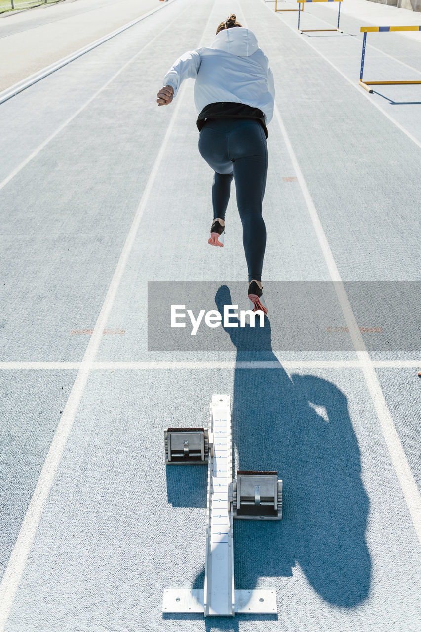 Back view of unrecognizable sportswoman beginning to run fast from starting blocks during track and field workout on stadium