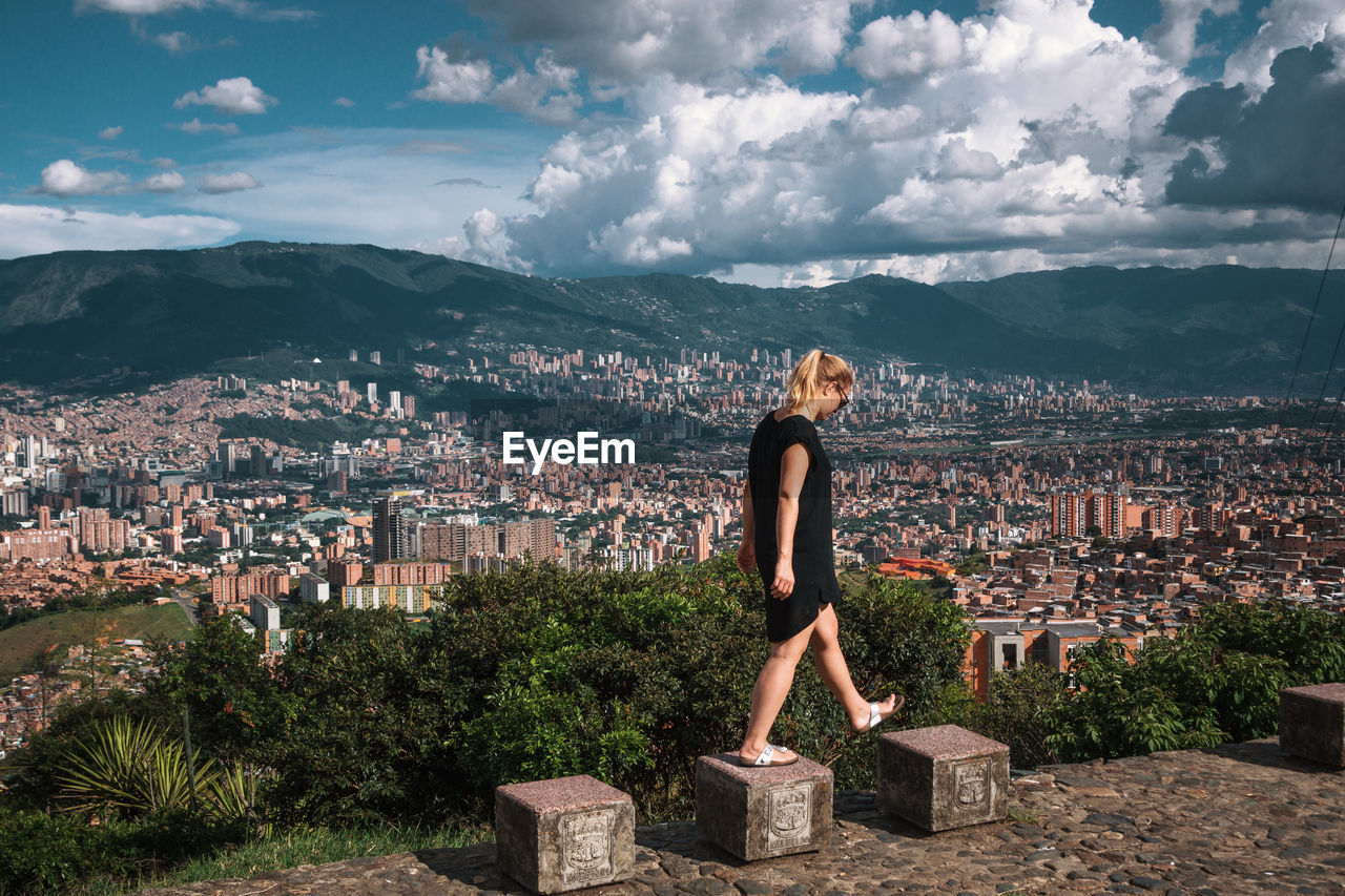 Side view of woman walking stone seats with buildings in backgrounds against cloudy sky