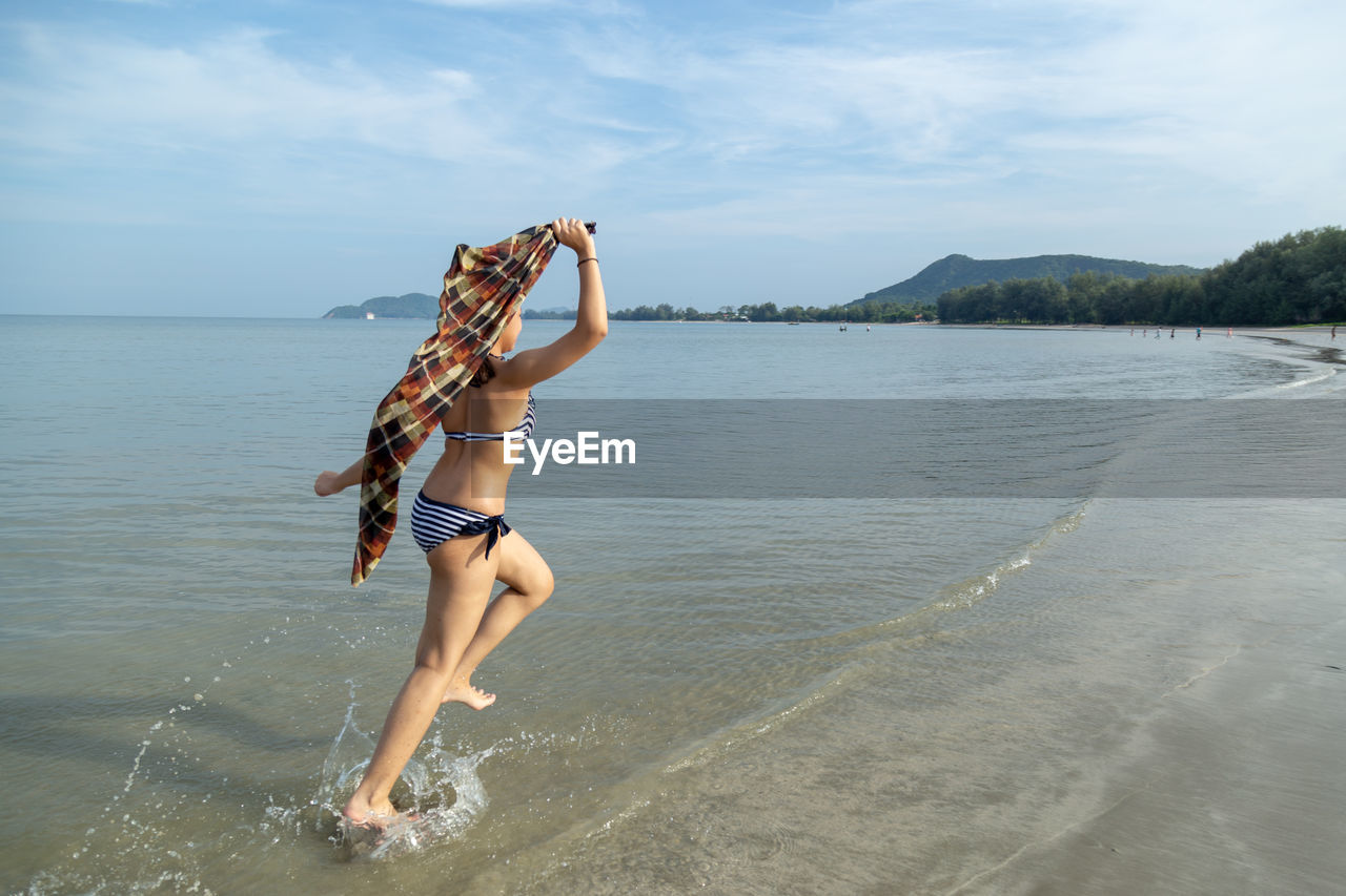 Teenage girl wearing bikini running at shore against sky
