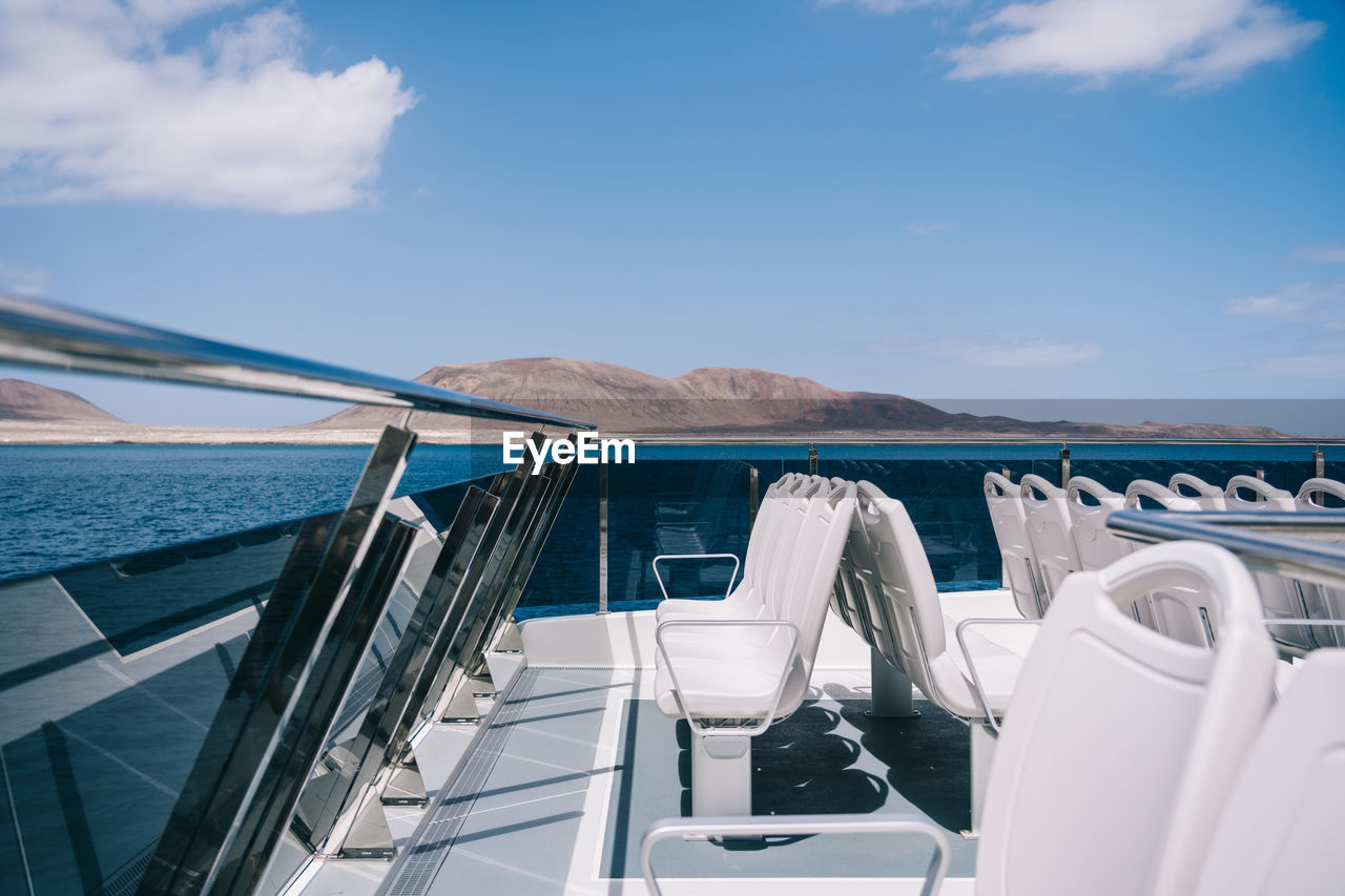 Empty white chairs on deck of cruise boat sailing in blue sea water with mountain on shore