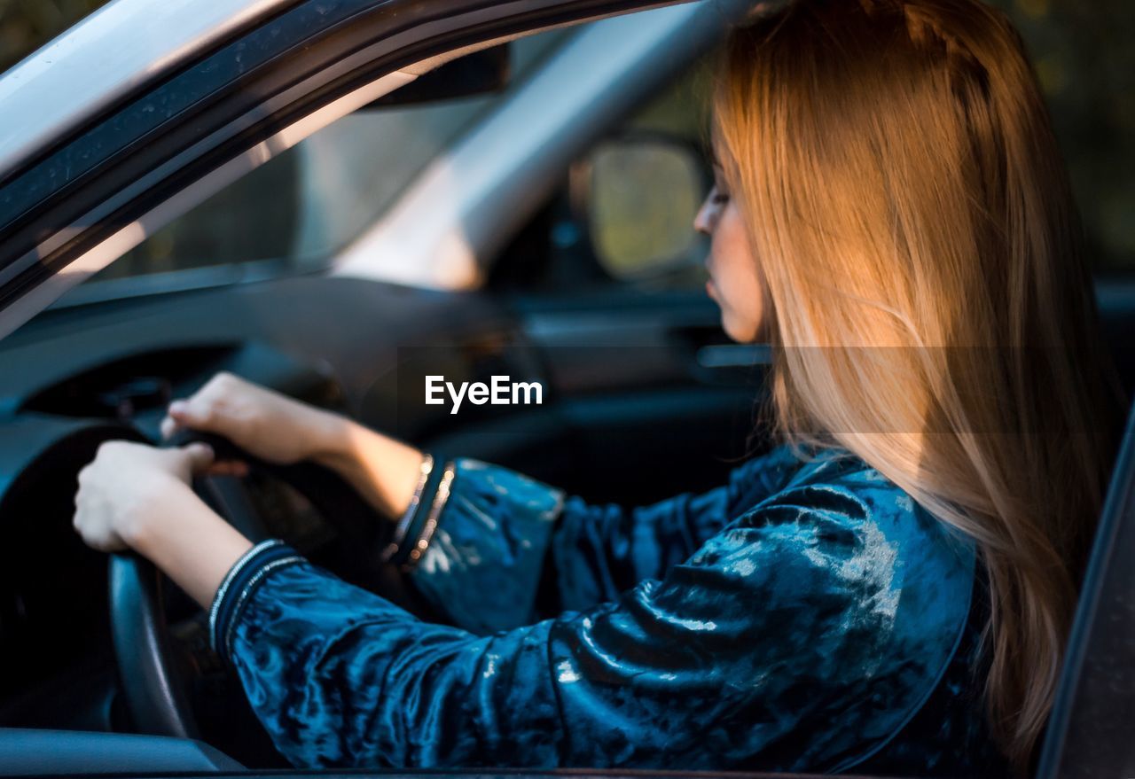 Side view of young woman with blond hair driving car seen through window