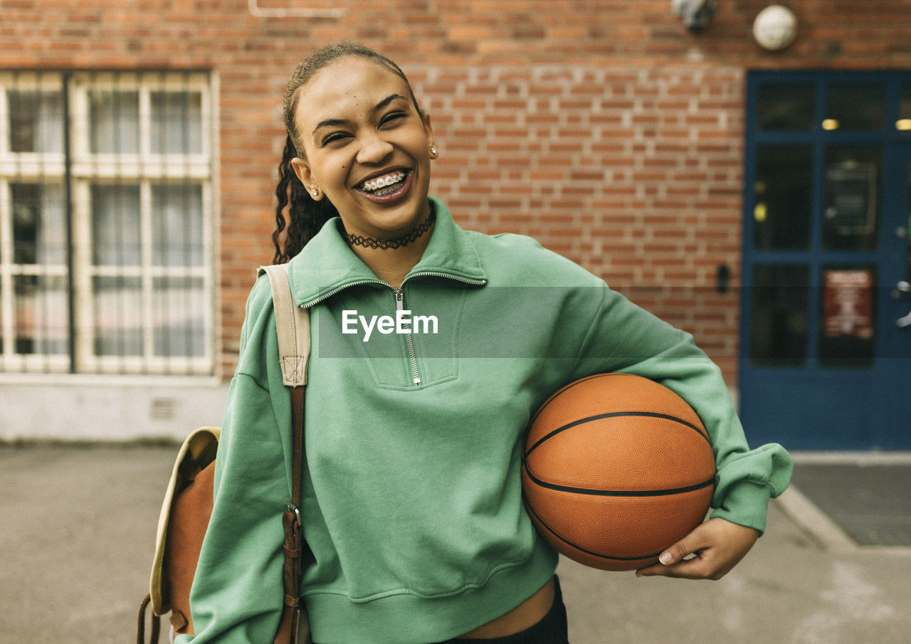 Portrait of happy young woman holding basketball under arm while standing in front of building