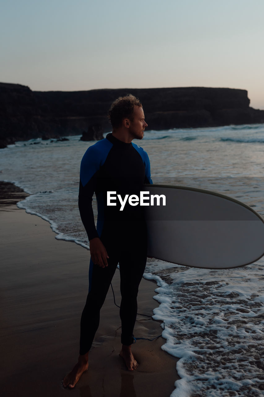 Full length of man in wetsuit with surfboard  standing on beach against sky