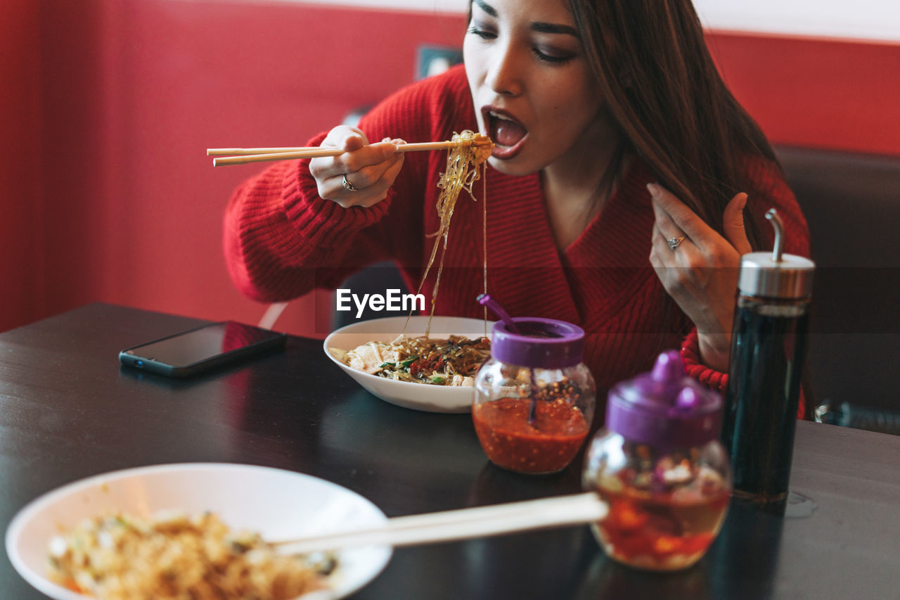Beautiful young asian woman in red clothes eating noodles with chopsticks in chinese restaurant