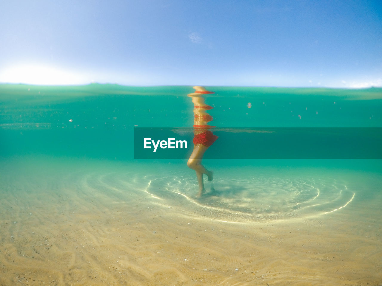 Underwater shot of female in red swimsuit in turquoise water standing on clear sandy bottom of lake