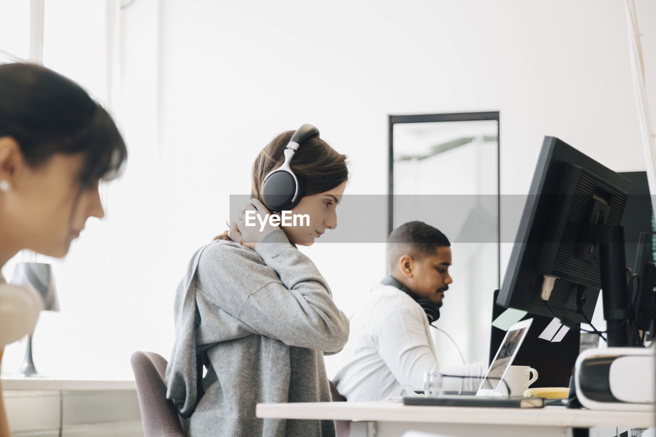 Focused female programmer with headphones using laptop on desk while sitting with coworkers in office