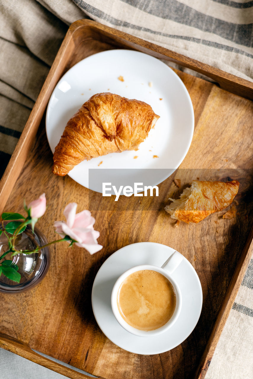 Fresh coffee with croissant on a wooden tray with a pink rose in a glass vase.