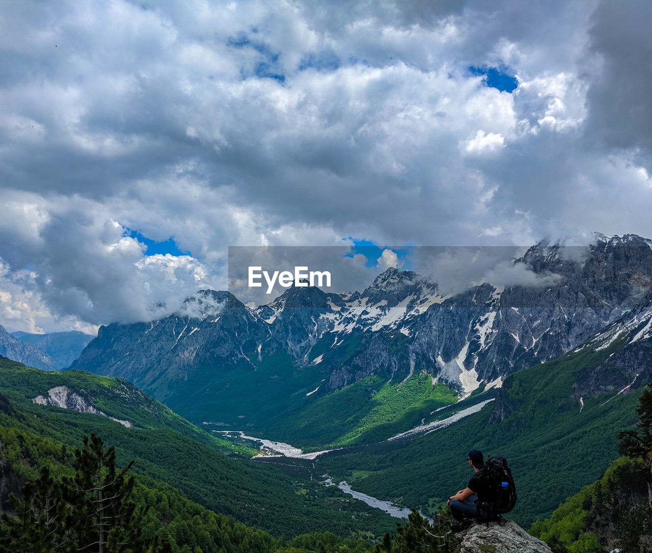 Side view of male hiker sitting on cliff with snowcapped mountains in background against cloudy sky