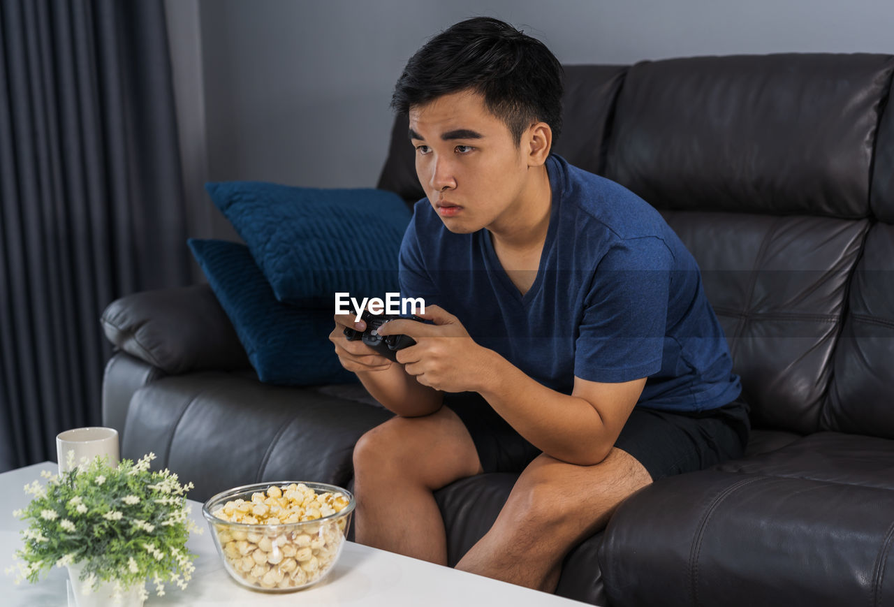 YOUNG MAN USING MOBILE PHONE WHILE SITTING ON SOFA AT HOME