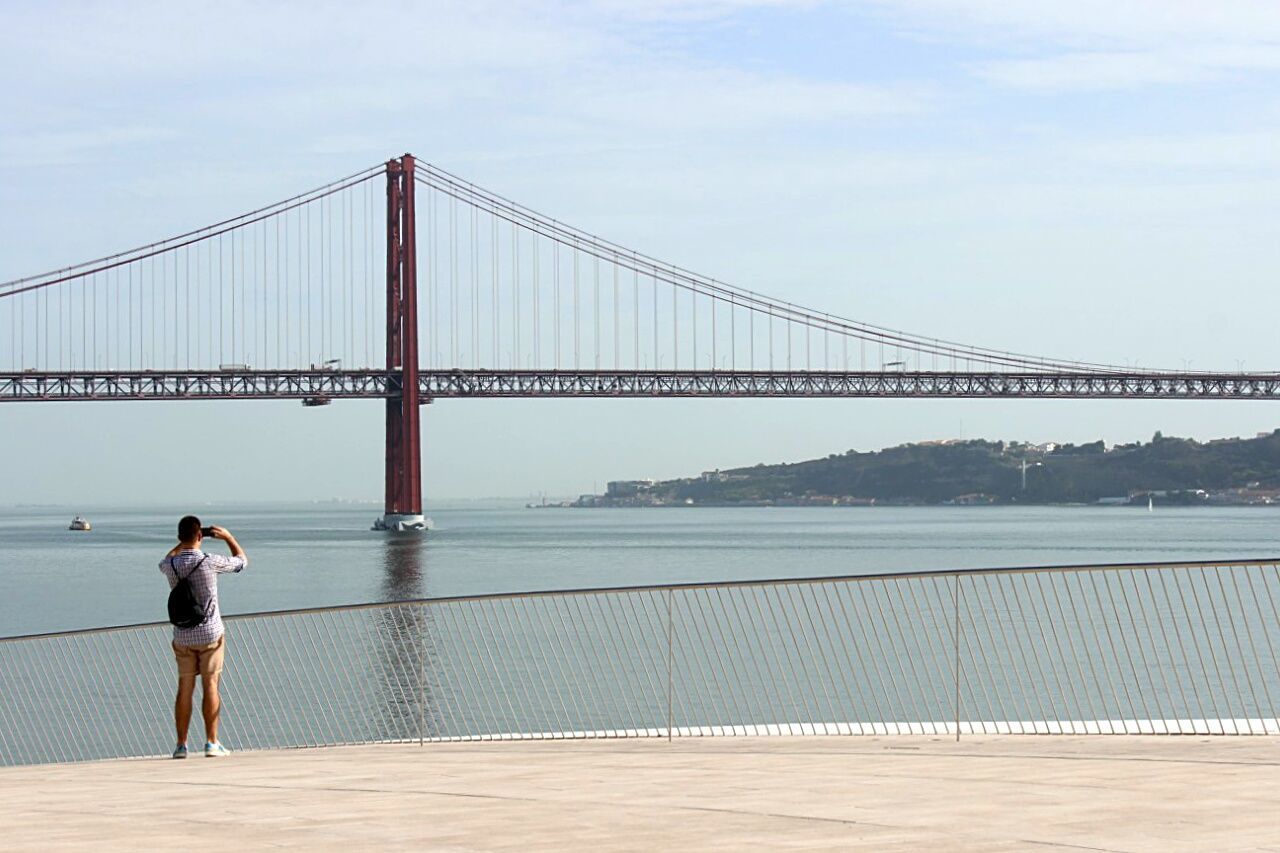 Rear view of man photographing 25 de abril bridge over tagus river against sky