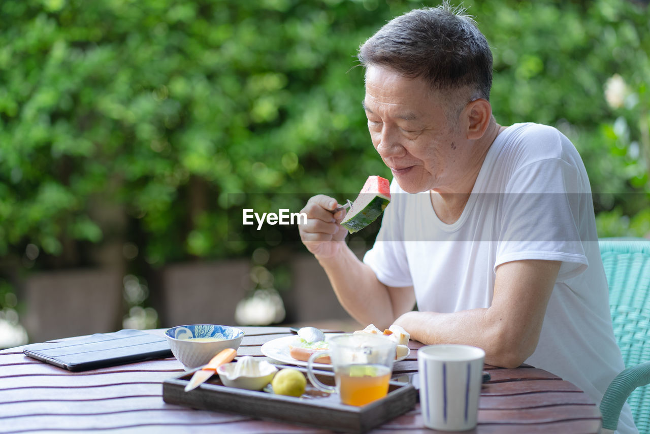 Smiling senior man eating watermelon while sitting on chair outdoors