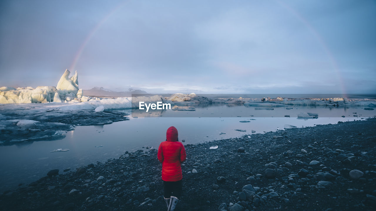 Rear view of woman by lake against cloudy sky during winter