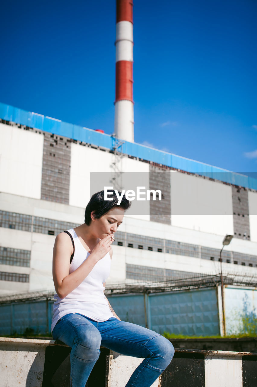 Young woman smoking cigarette while sitting on barricade against factory