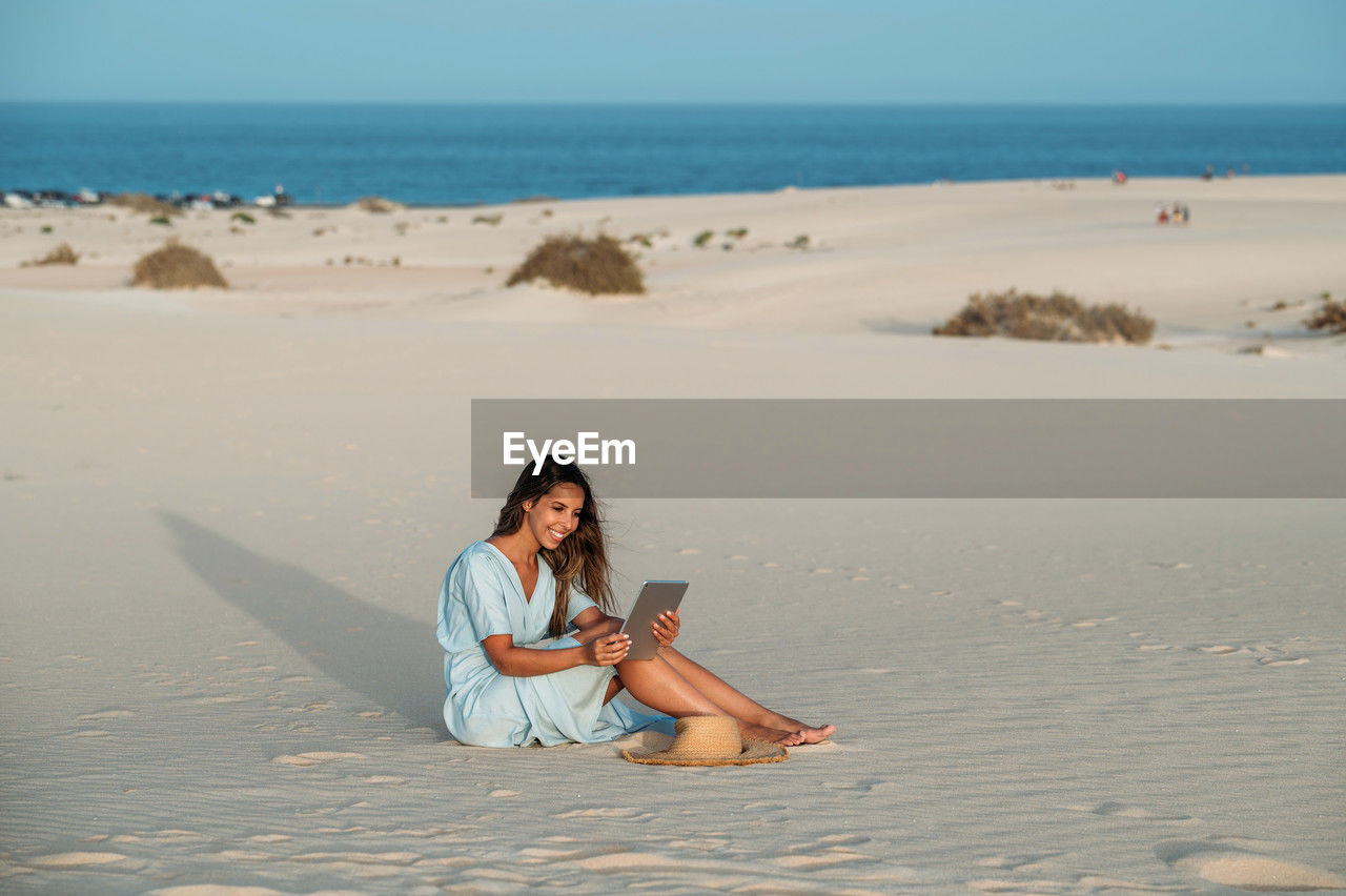 Sincere young barefoot female tourist with tablet sitting on sandy shore against sea and dunes of corralejo in sunlight in fuerteventura canary islands spain