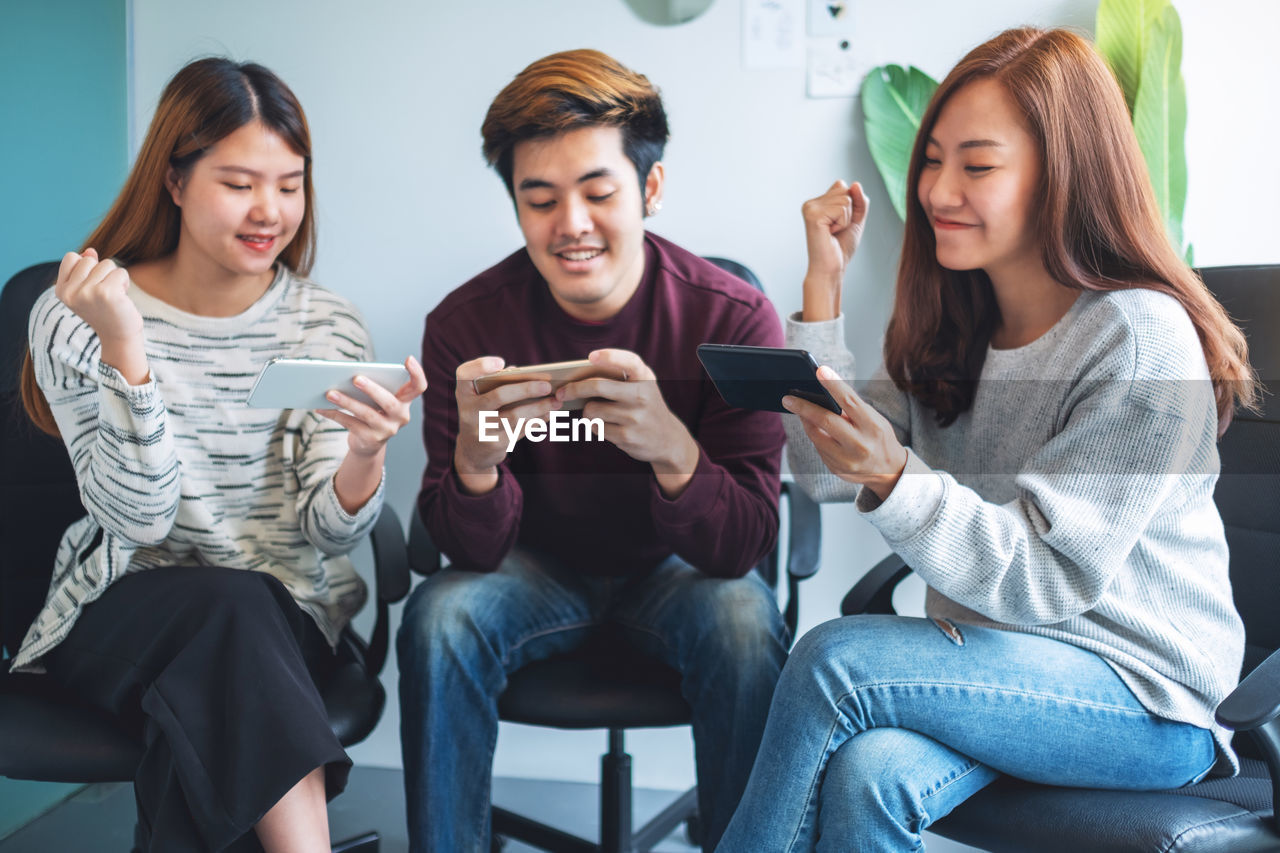 smiling friends using smart phones while sitting on sofa at school