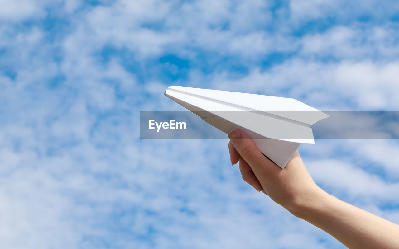 Cropped hand of woman playing with paper airplane against cloudy sky