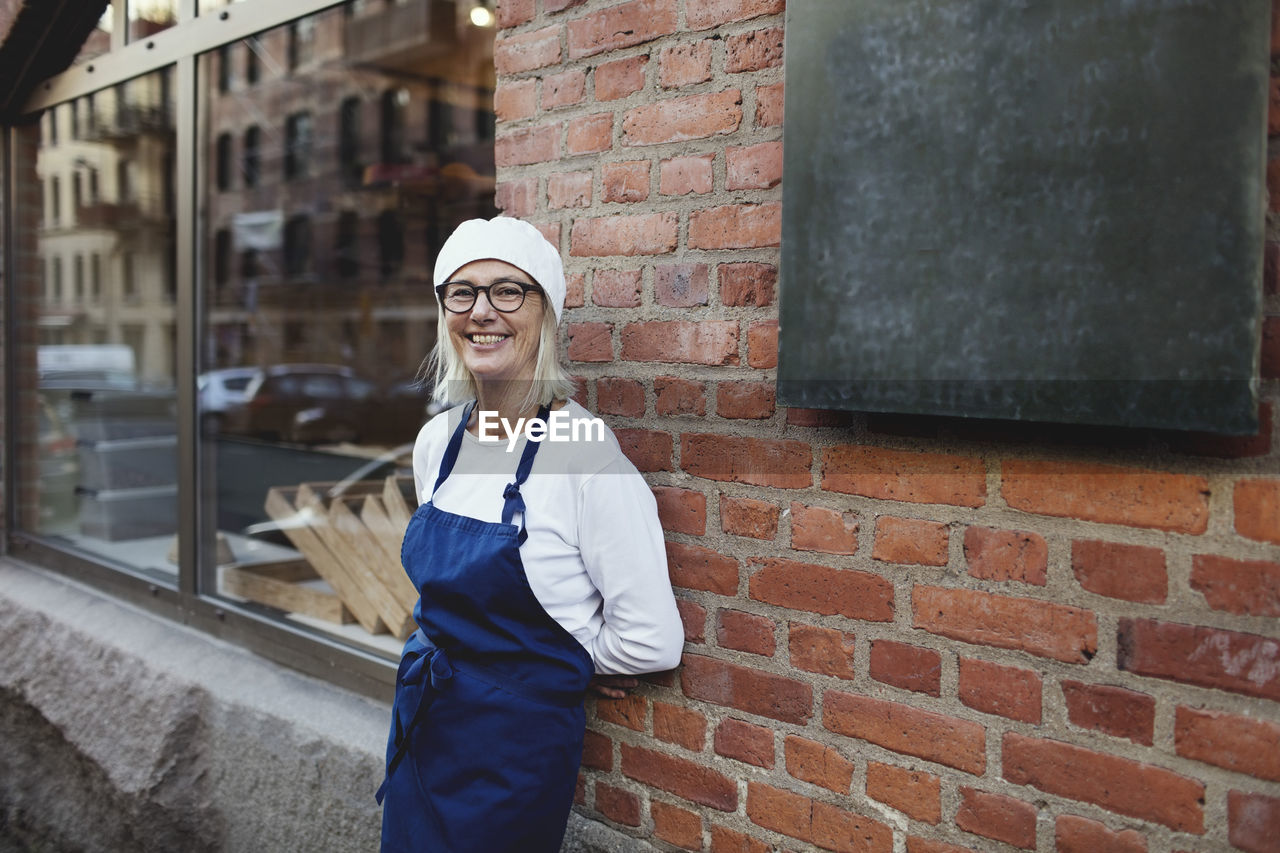 Portrait of smiling senior baker leaning on brick wall outside bakery