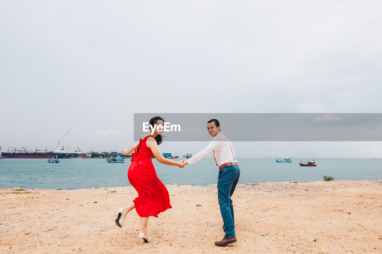 Rear view of couple holding hands while standing on beach against sky