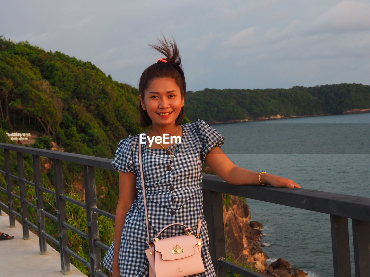 Portrait of smiling young woman standing by railing against sky