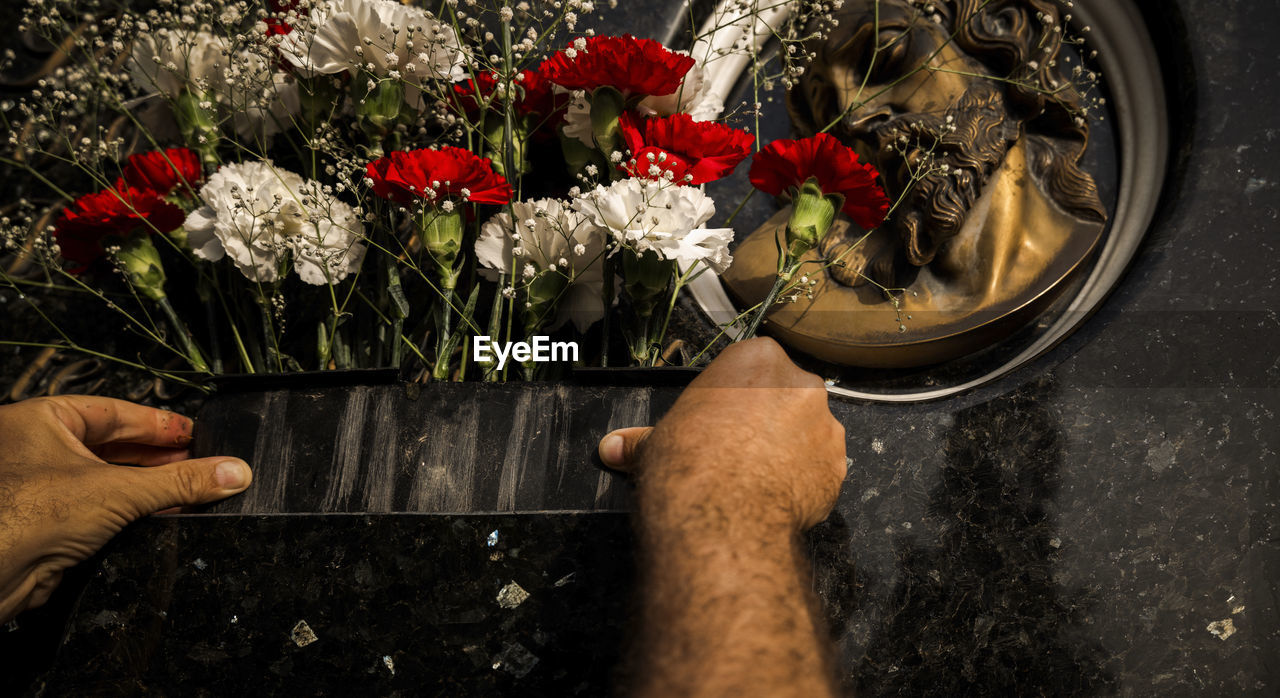 Close-up of hands offering bouquet of red and white flowers in cemetery