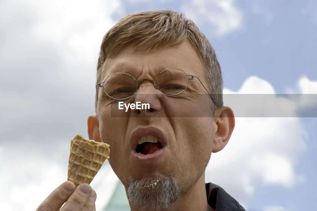 Low angle portrait of mature man making face while having ice cream cone against sky