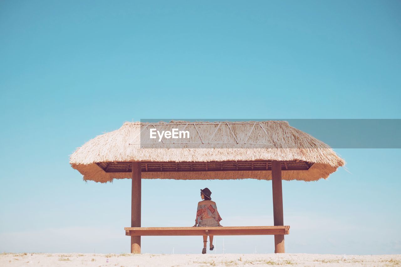 Rear view of woman sitting on seat below thatched roof against blue sky