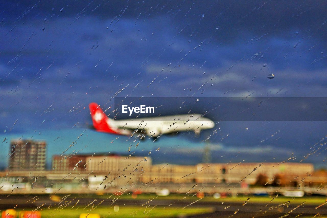 AIRPLANE FLYING AGAINST CLOUDY SKY SEEN THROUGH WET WINDOW