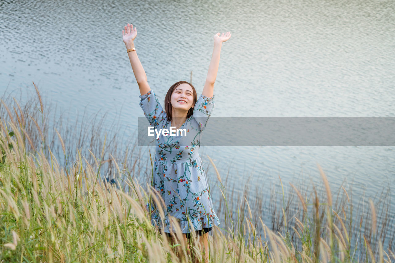Smiling young woman with arms raised standing by lake