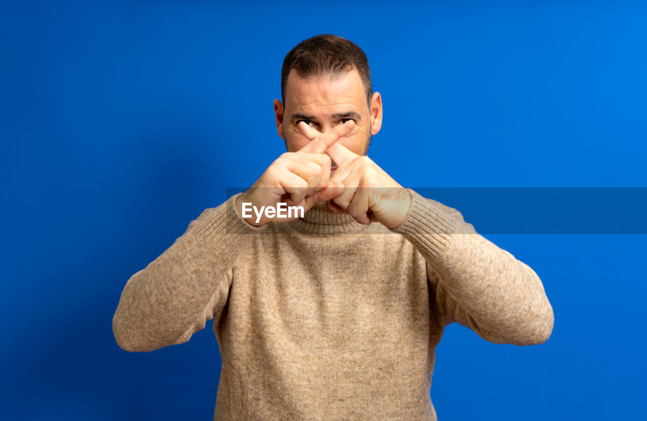 one person, blue, finger, men, studio shot, adult, colored background, waist up, blue background, hand, person, front view, portrait, emotion, arm, indoors, human face, negative emotion, gesturing, standing, nose, sign language, facial hair, clothing, copy space, anger, casual clothing