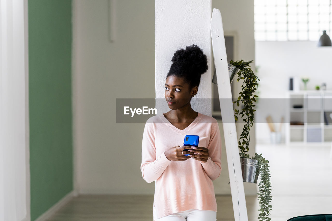 Thoughtful woman with mobile phone looking away while standing at home