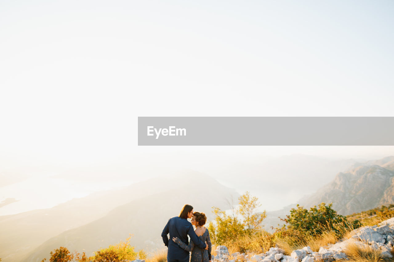 REAR VIEW OF COUPLE KISSING AGAINST MOUNTAIN