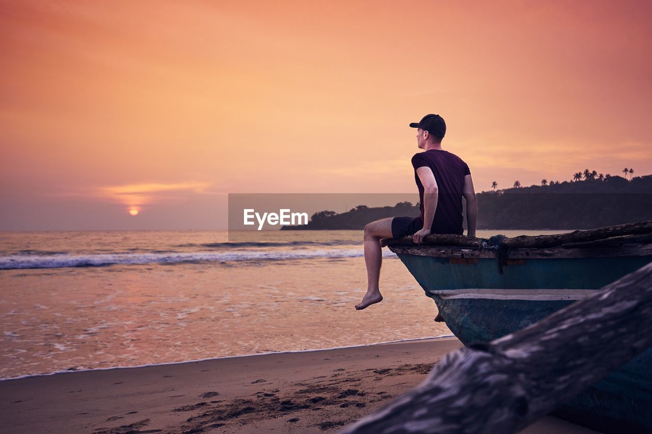 Man sitting on boat moored at beach against sky during sunset